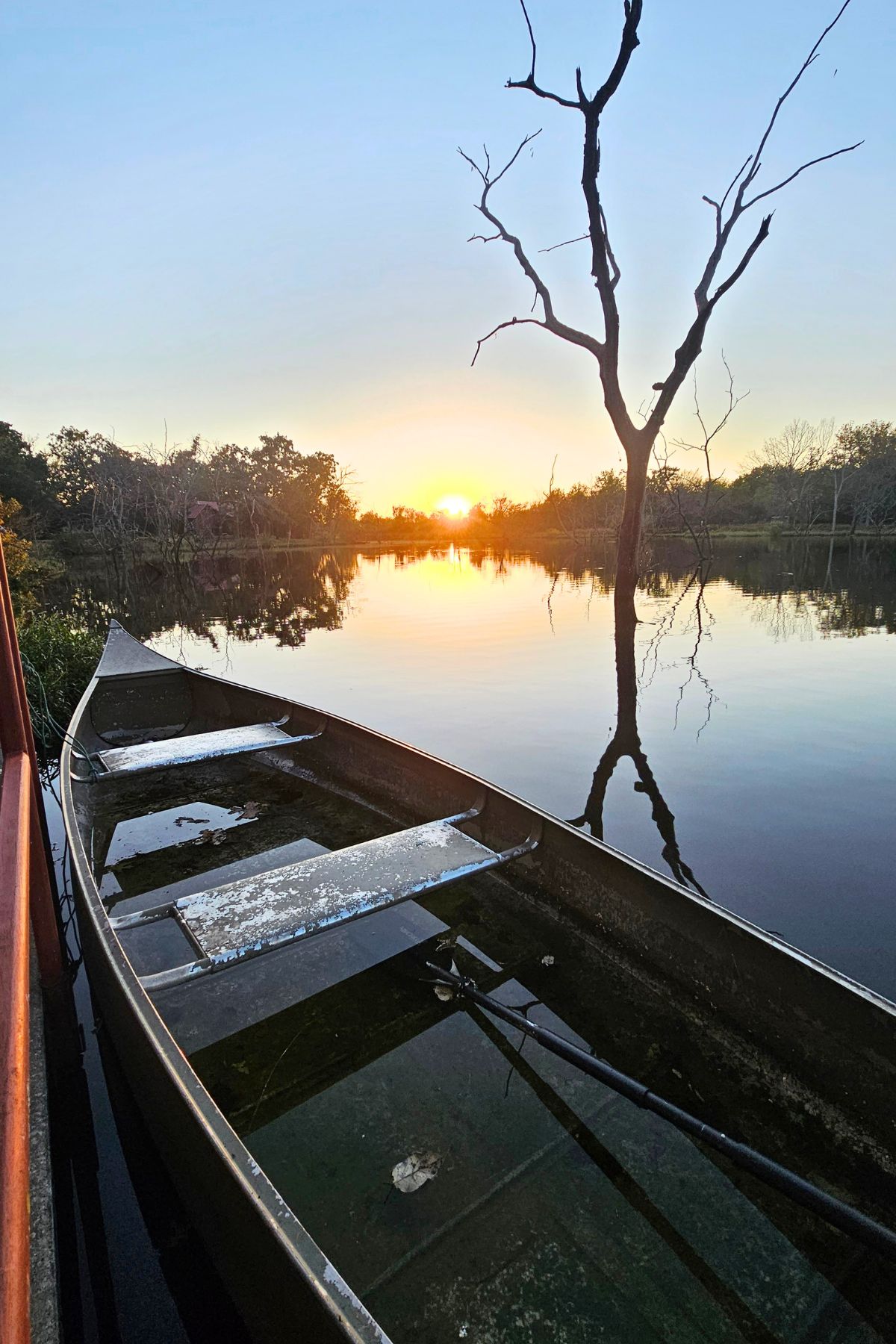 A partially sunken wooden boat rests in calm water at sunset in Southeast Kansas, with a leafless tree and forest reflected in the lake.
