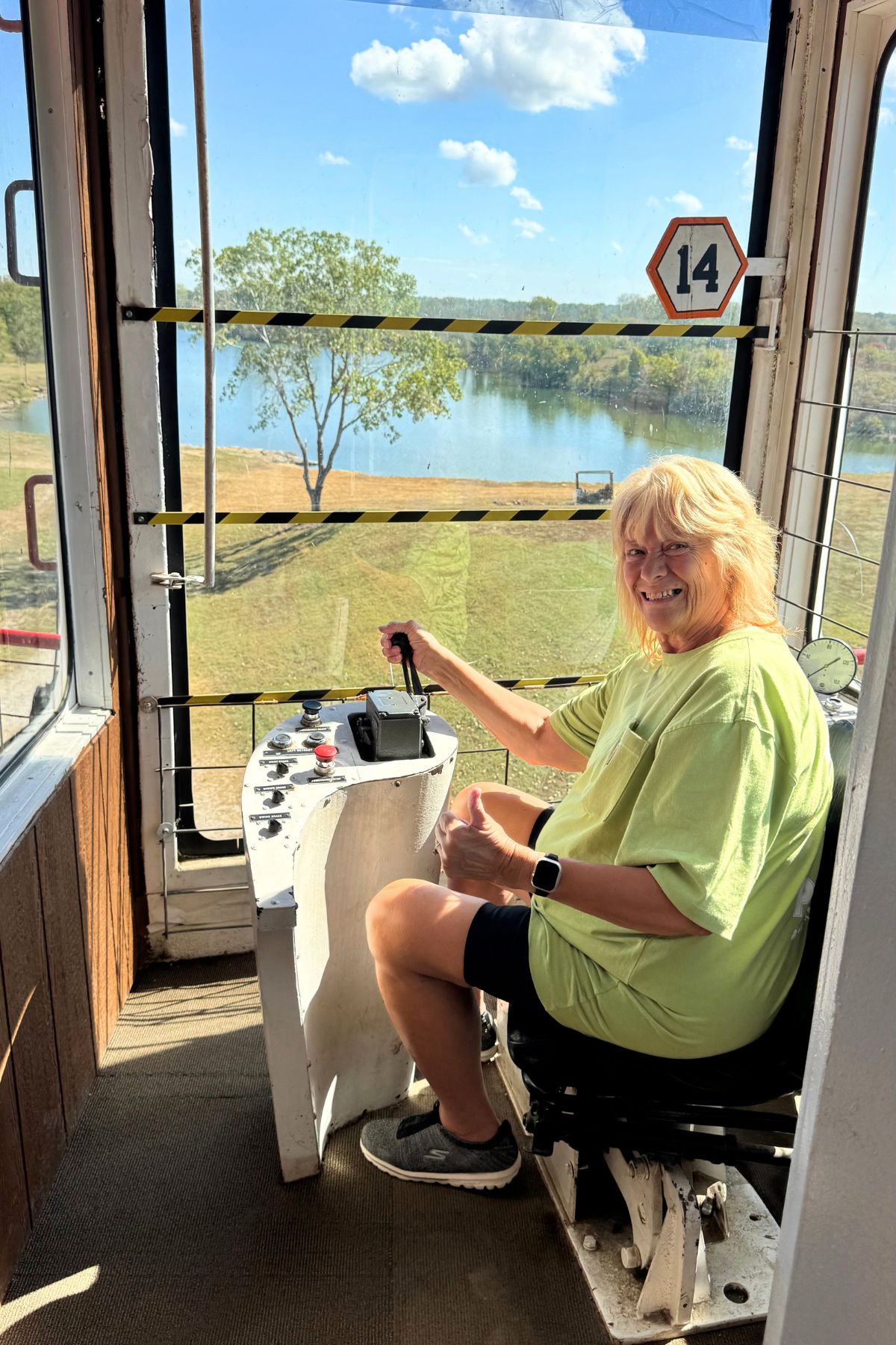 A woman in a green shirt sits at the controls of a small booth with windows, giving a thumbs up. A lake and tree are visible outside on a sunny Southeast Kansas day.