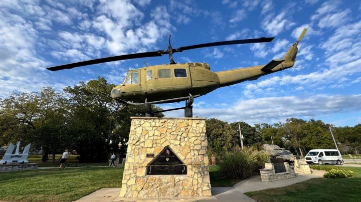 A military helicopter mounted on a stone pedestal serves as a memorial in a park in Southeast Kansas, surrounded by trees, grass, and a partly cloudy sky.