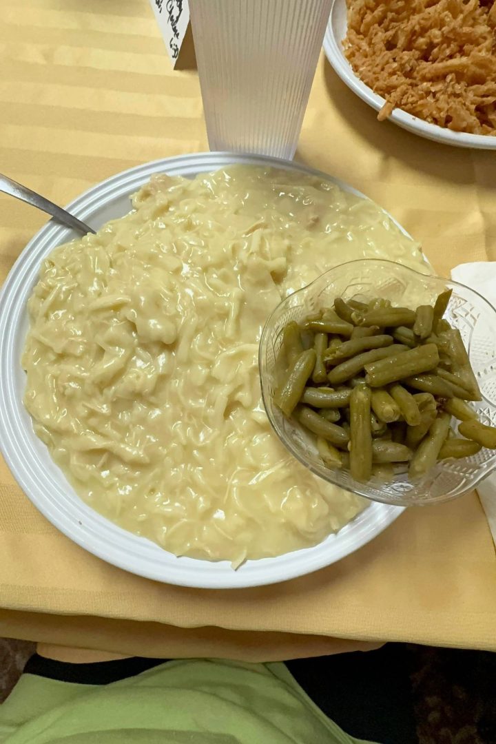 A plate with creamy noodles and a side of green beans in a glass bowl, set with a fork and clear plastic cup on a yellow tablecloth, evokes the comforting flavors of Southeast Kansas home cooking.