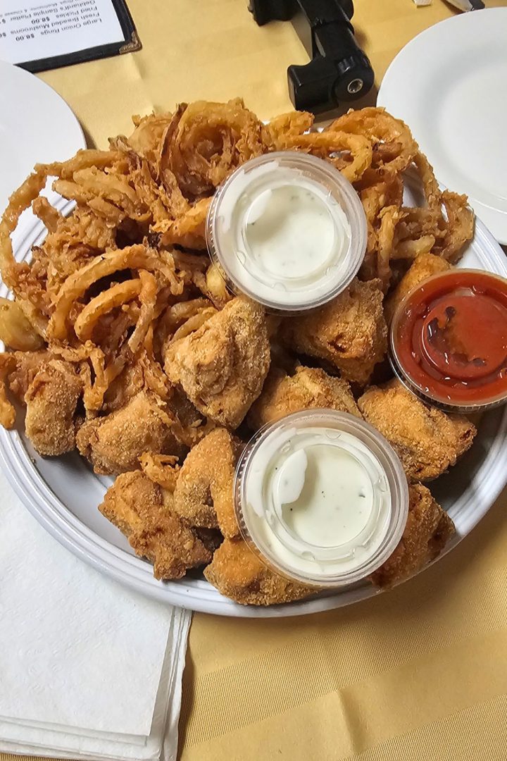 A plate of fried onion rings and breaded chicken pieces, inspired by Southeast Kansas flavors, is served with two cups of ranch dip and one cup of red sauce on a table.
