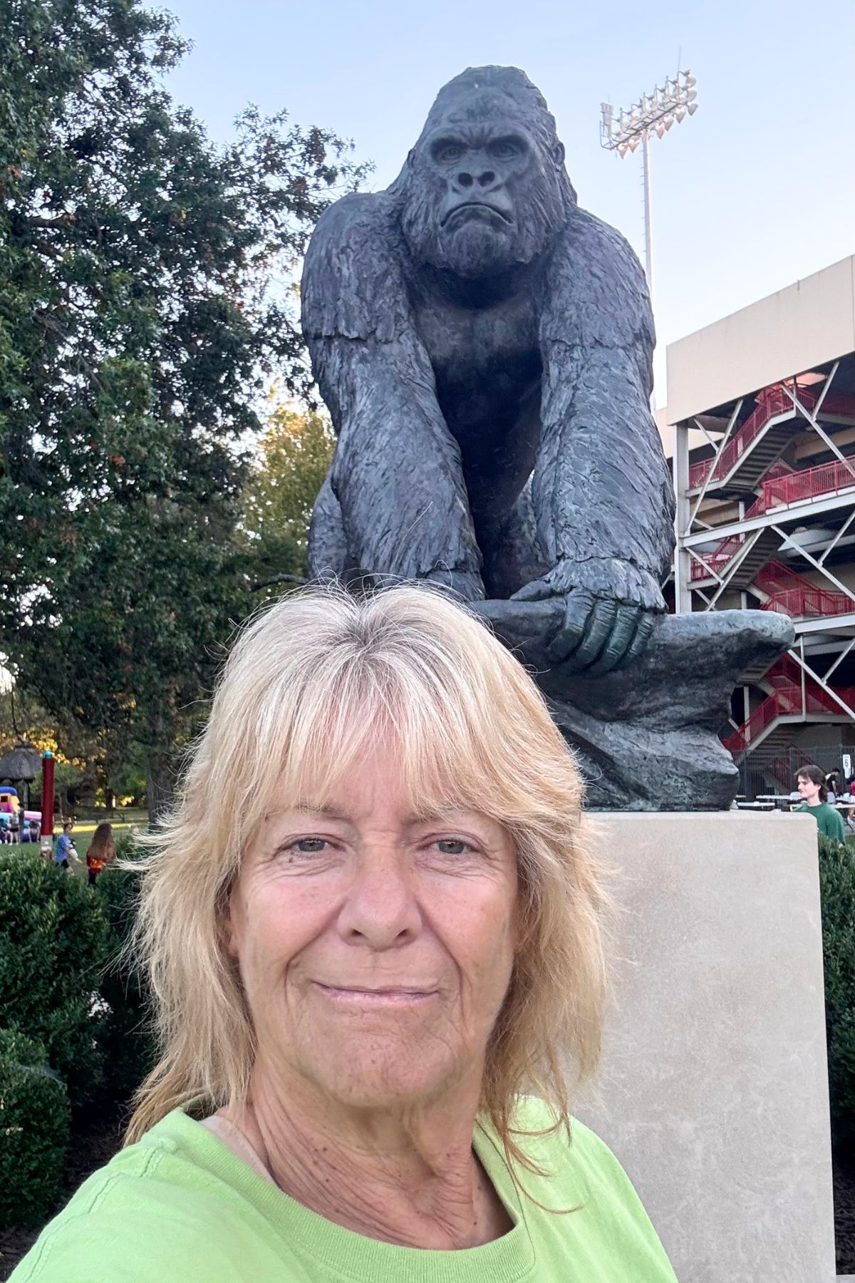 A woman takes a selfie in front of a large gorilla statue outdoors in Southeast Kansas, with trees and a stadium structure in the background.
