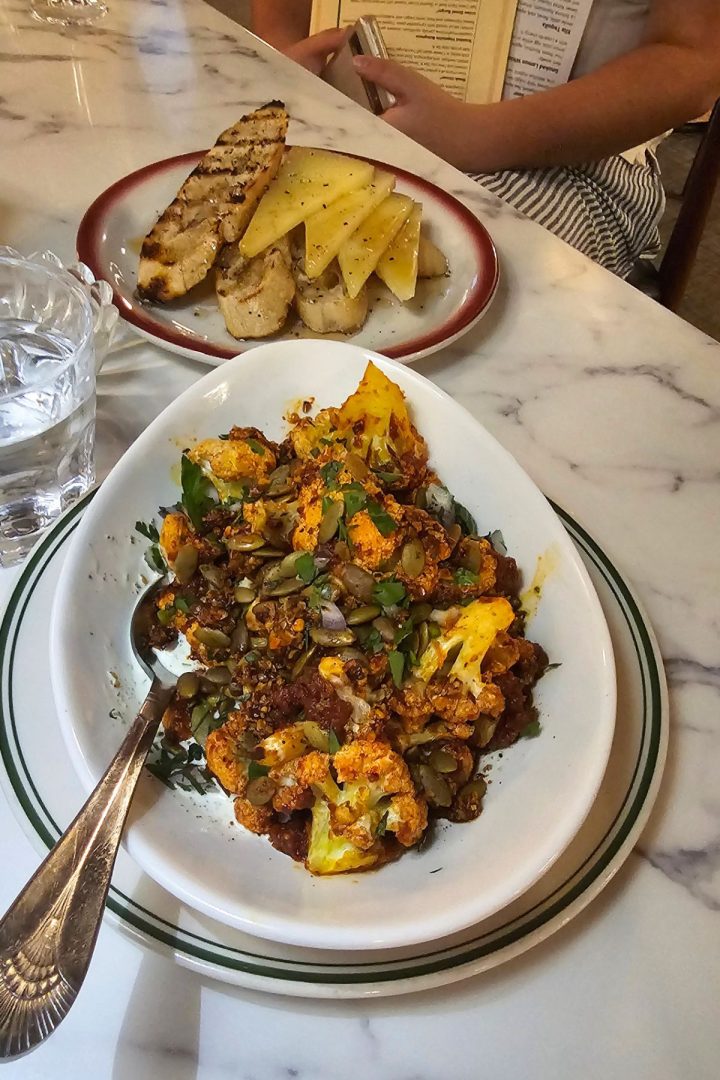 A white bowl of roasted cauliflower with seeds and herbs sits beside a plate of grilled bread and sliced cheese on a marble table, inspired by the fresh flavors found in Southeast Kansas.