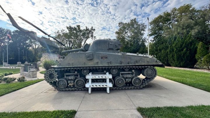 A military tank is displayed outdoors on a concrete pad in Southeast Kansas, surrounded by grass and trees, with a white step ladder placed in front of it.