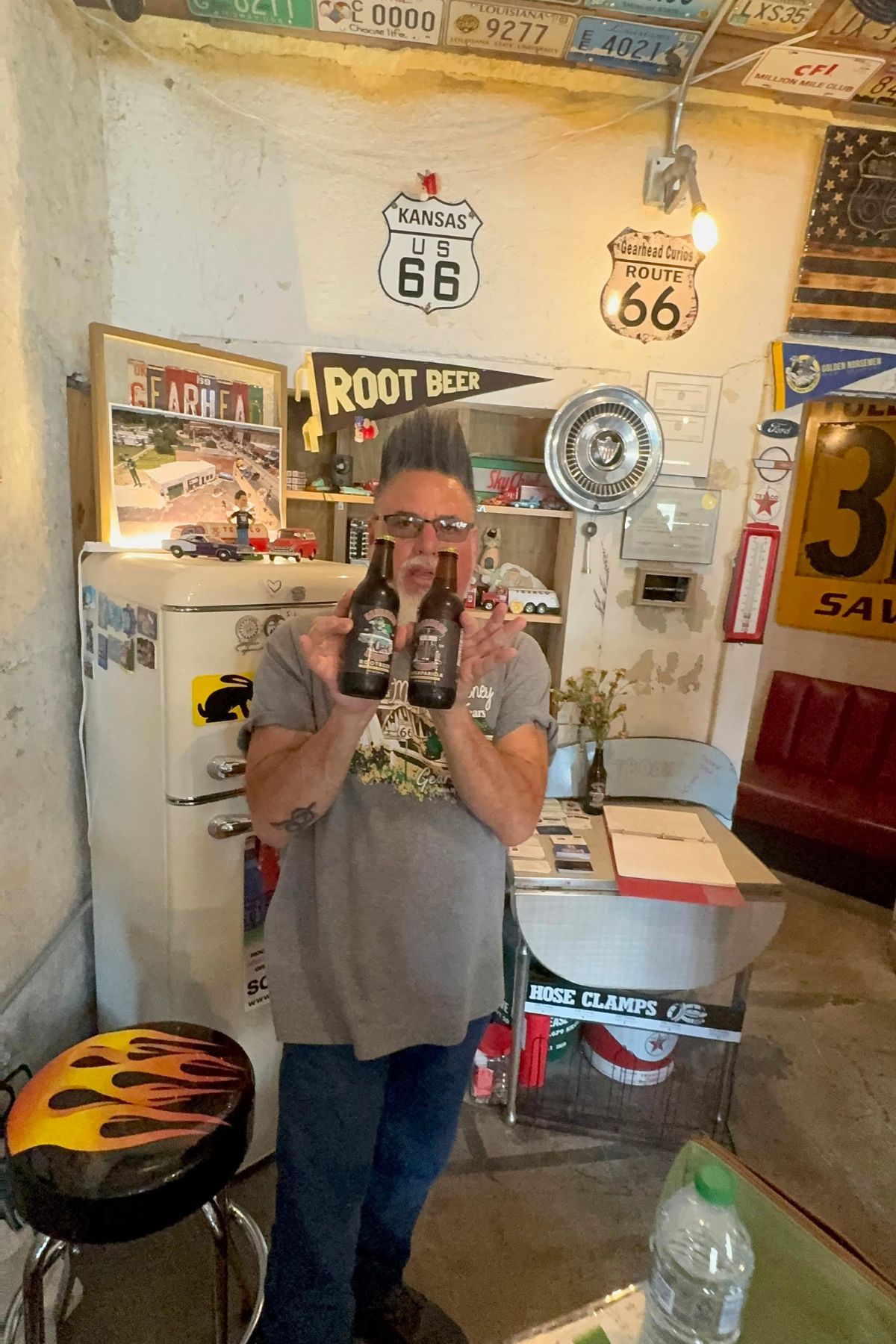 A man stands in a retro-themed room holding two glass bottles, with vintage signs, a fridge, and Southeast Kansas memorabilia visible in the background.