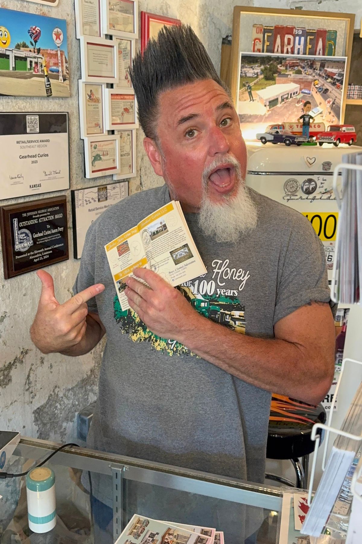 A man with styled hair and a gray beard stands behind a counter in Southeast Kansas, smiling and pointing at a brochure he is holding in one hand.