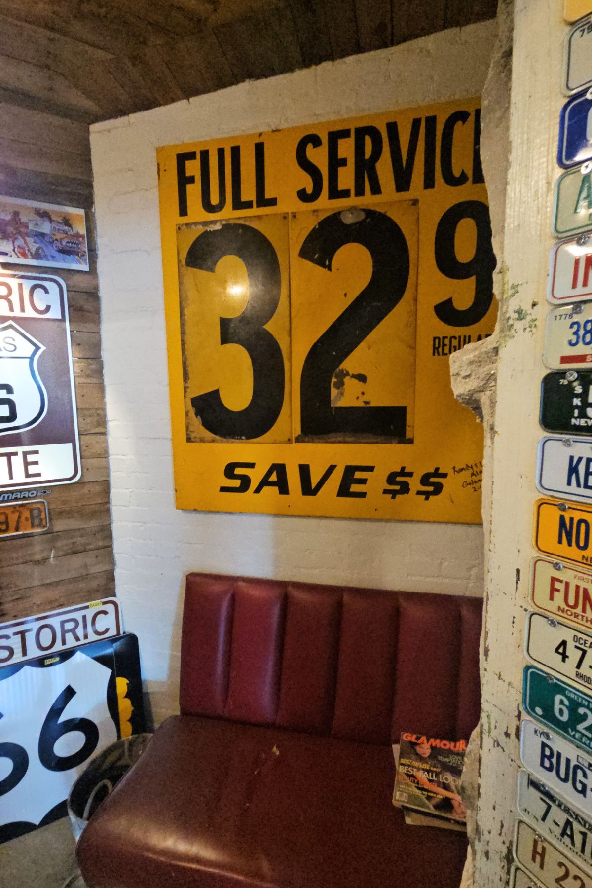 A vintage yellow gas station sign reading "FULL SERVICE 32.9 SAVE $$" hangs above a red booth seat, surrounded by old license plates on the walls—a nostalgic scene straight out of Southeast Kansas.