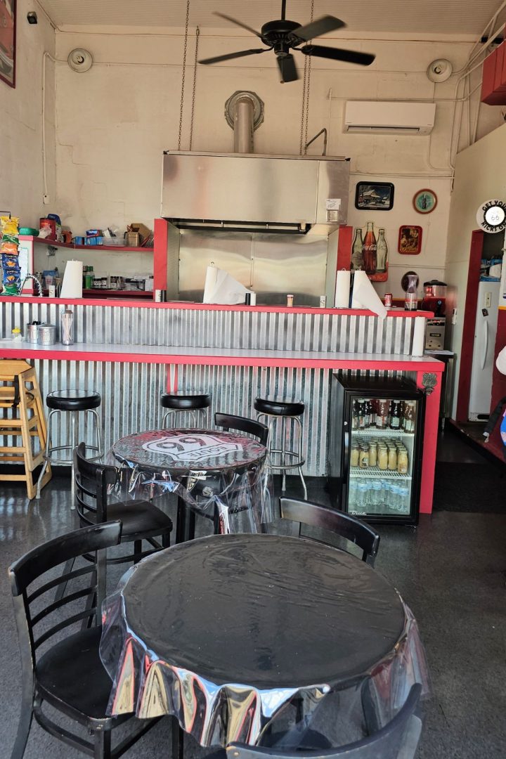 Small Southeast Kansas diner with black tables and chairs, a corrugated metal counter, barstools, a ceiling fan, and a mini fridge below the counter; shelves with snacks are visible in the background.