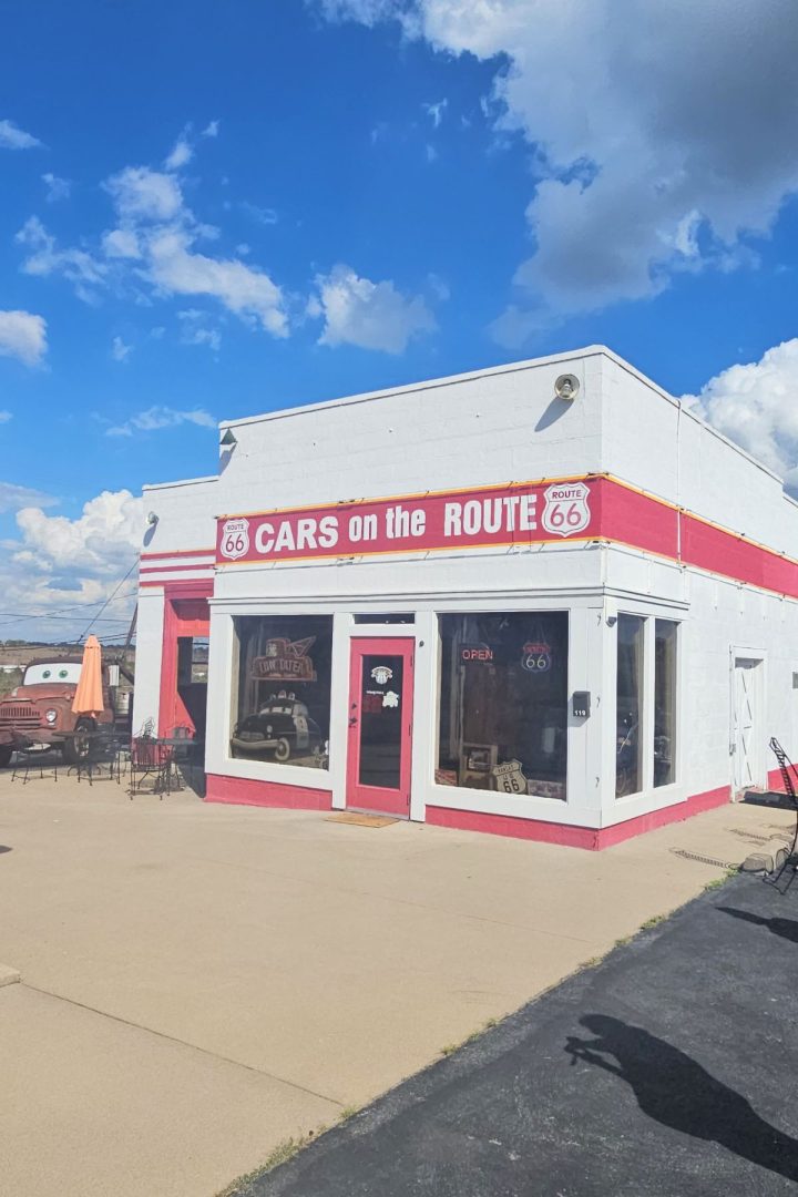 A white and red building with a sign reading "Cars on the Route" on Route 66 in Southeast Kansas, outdoor seating, and a clear blue sky with clouds above.