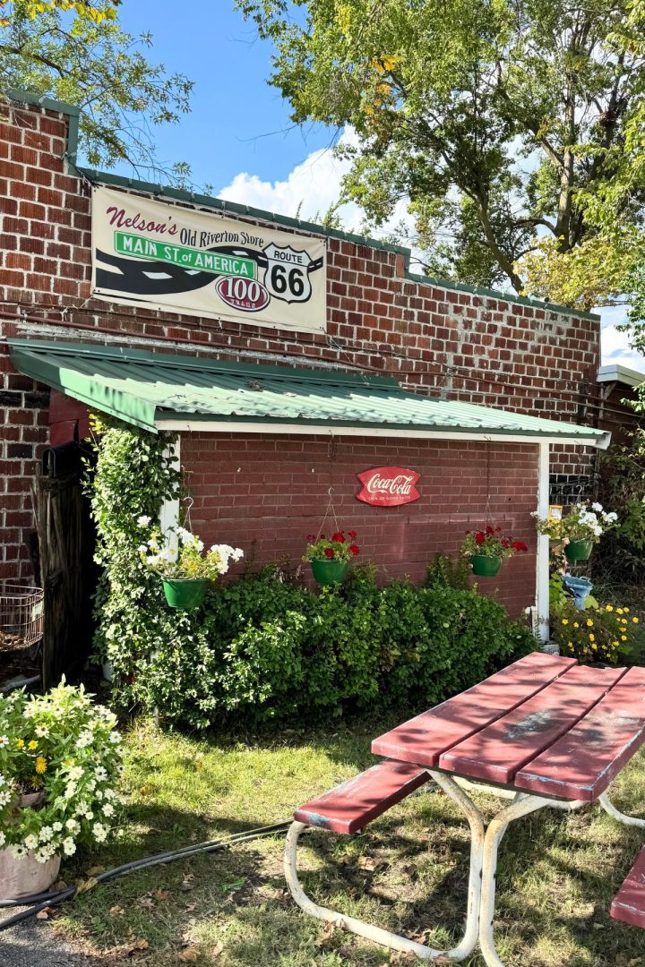 A brick building with a green awning in Southeast Kansas features a Route 66 sign, potted flowers, and a Coca-Cola sign above a picnic table out front.