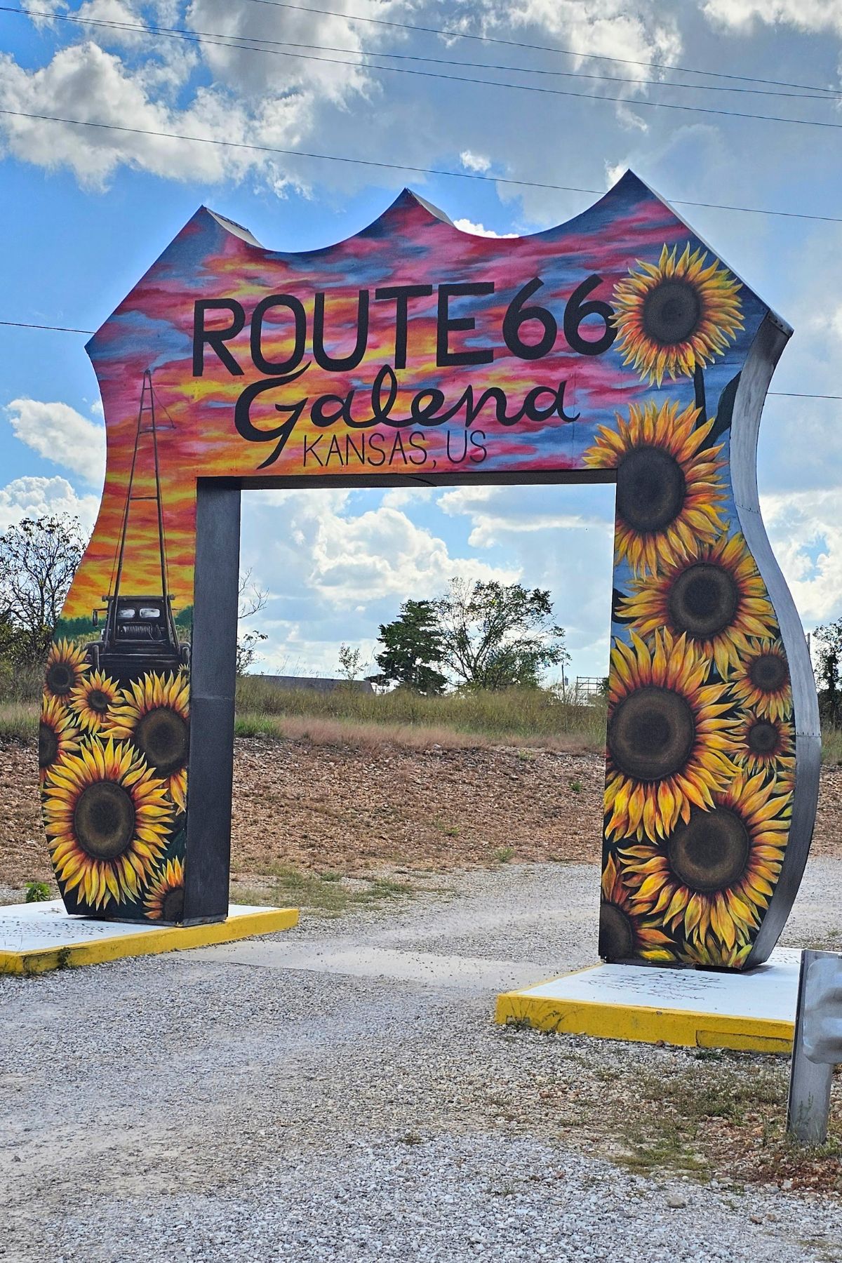 A large Route 66 sign-shaped archway in Galena, Southeast Kansas, decorated with sunflowers, a car, and a sunset sky.