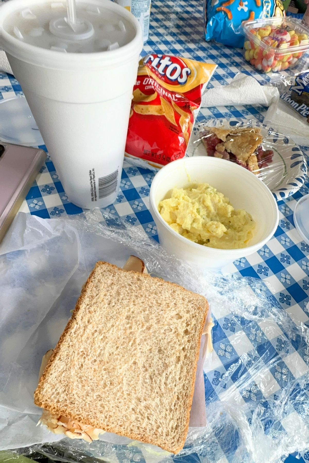 A classic Southeast Kansas lunch: a turkey sandwich on wheat bread, a cup of potato salad, Fritos, a large drink, and other snacks arranged on a blue and white checkered tablecloth.