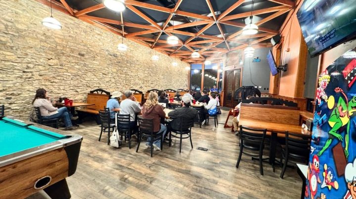 People sit at tables in a casual Southeast Kansas restaurant with stone walls, wooden beams, a pool table, and a colorful arcade machine in the foreground.