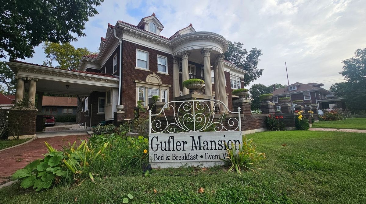 Large brick mansion with columns and a sign in front reading "Gulfer Mansion Bed & Breakfast • Event Venue," surrounded by grass and flowers on a cloudy day in Southeast Kansas.