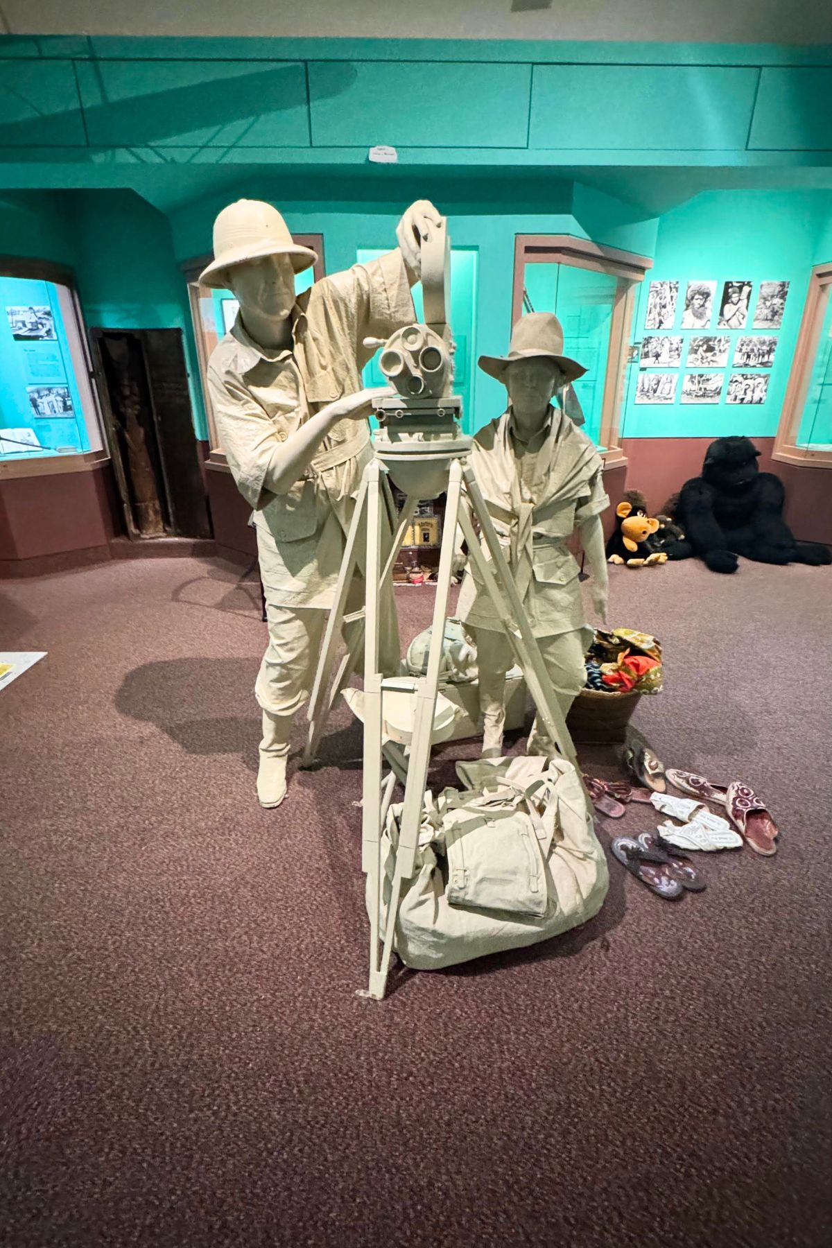 Two mannequins dressed as explorers stand by a surveying instrument and tripod; travel bags, hats, and shoes are on the floor nearby in this Southeast Kansas museum exhibit.