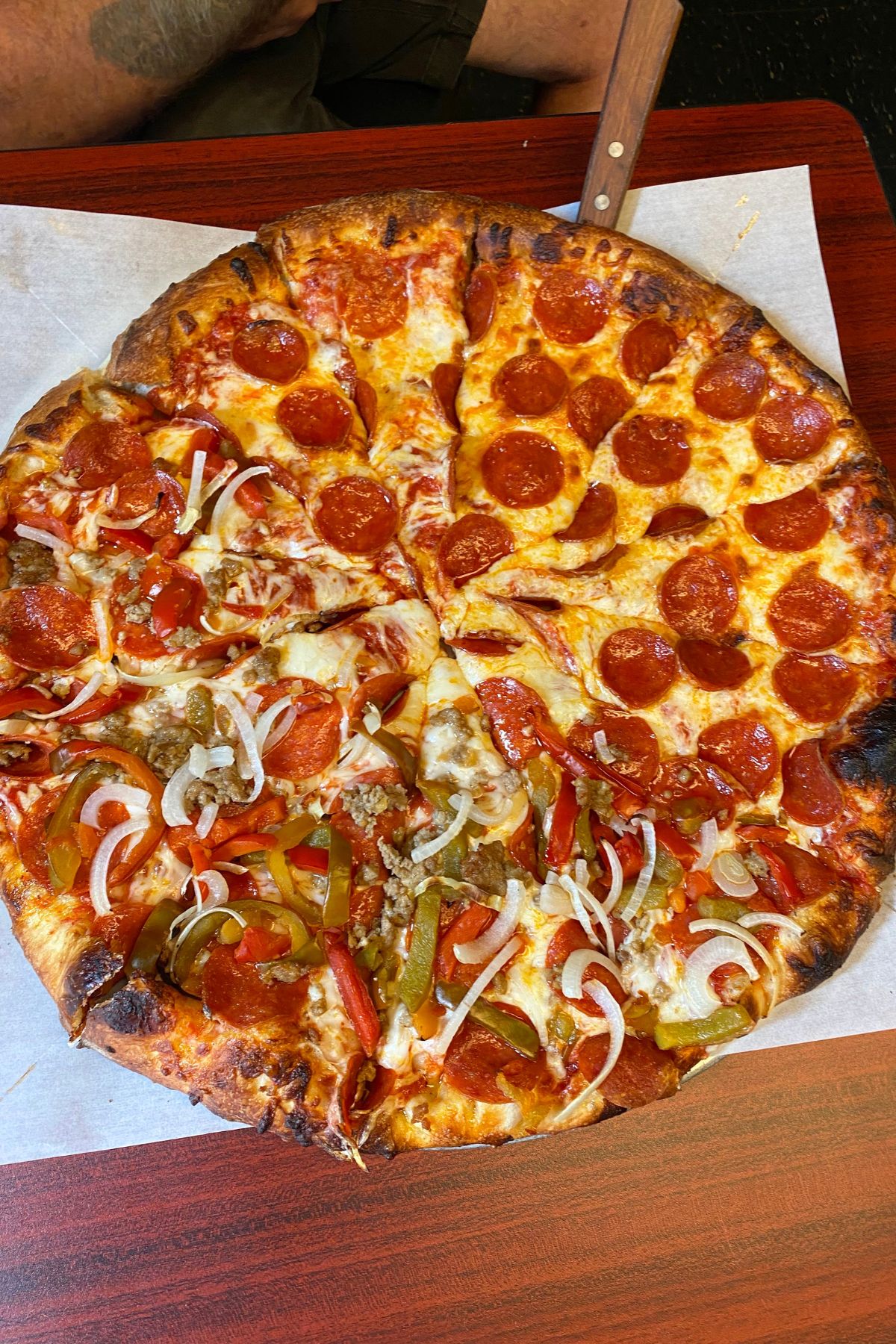 A large pizza on a wooden table, half topped with pepperoni, and half with pepperoni, sausage, green peppers, onions, and red peppers—perfect for sharing in Southeast Kansas. A hand and knife are visible in the background.