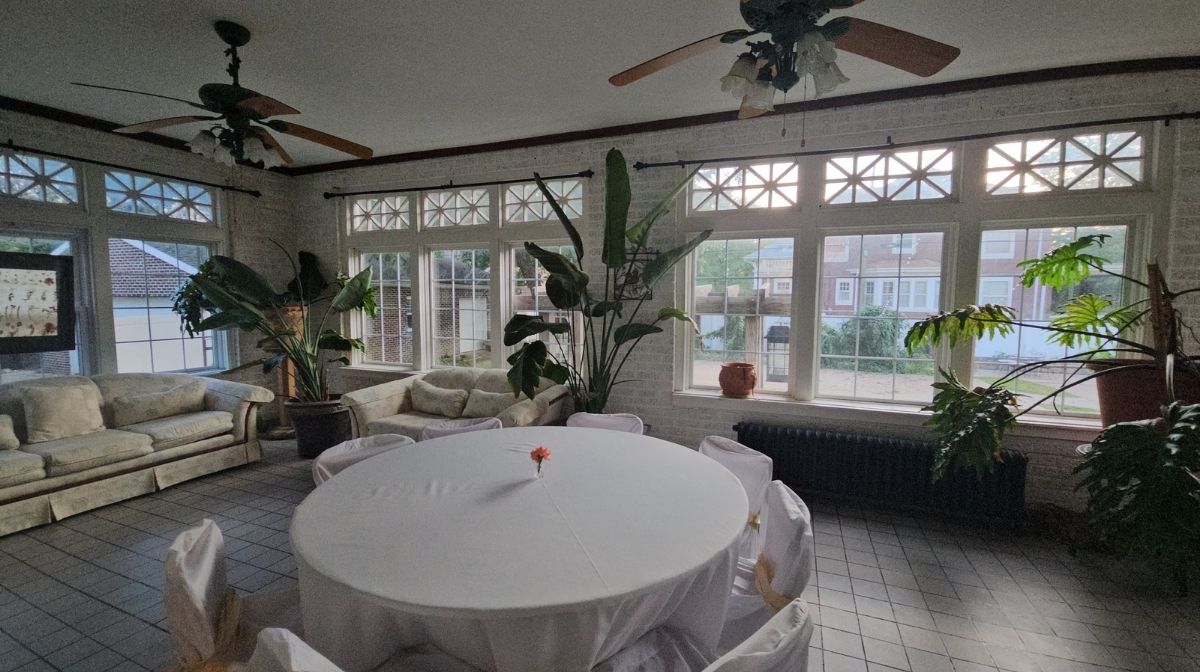 A bright room in Southeast Kansas with large windows, tile floor, ceiling fans, potted plants, a round table draped in white cloth, and white sofas lining the walls.
