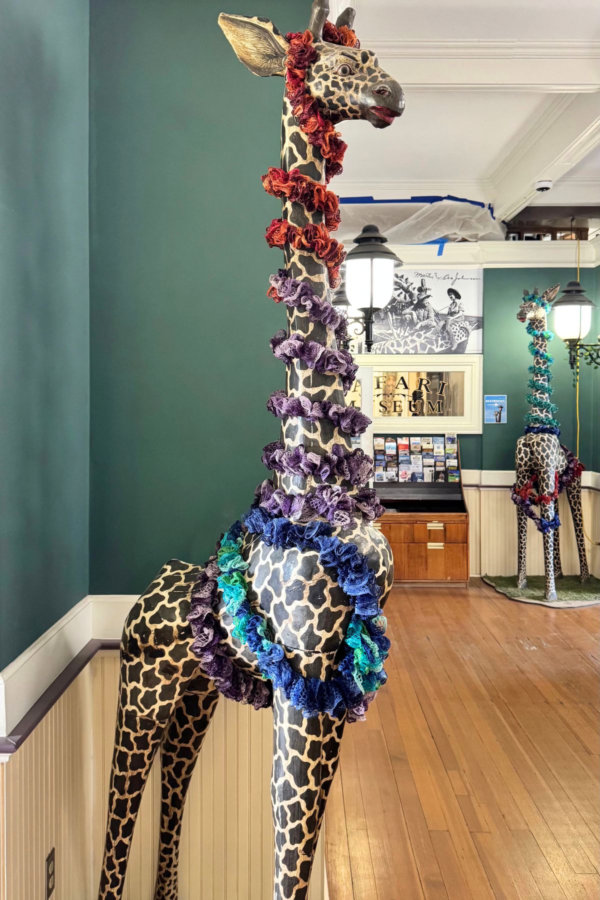 A life-sized giraffe statue decorated with multicolored ruffled fabric stands indoors on a wooden floor, bringing a touch of whimsical art to Southeast Kansas, with another similarly decorated giraffe in the background.