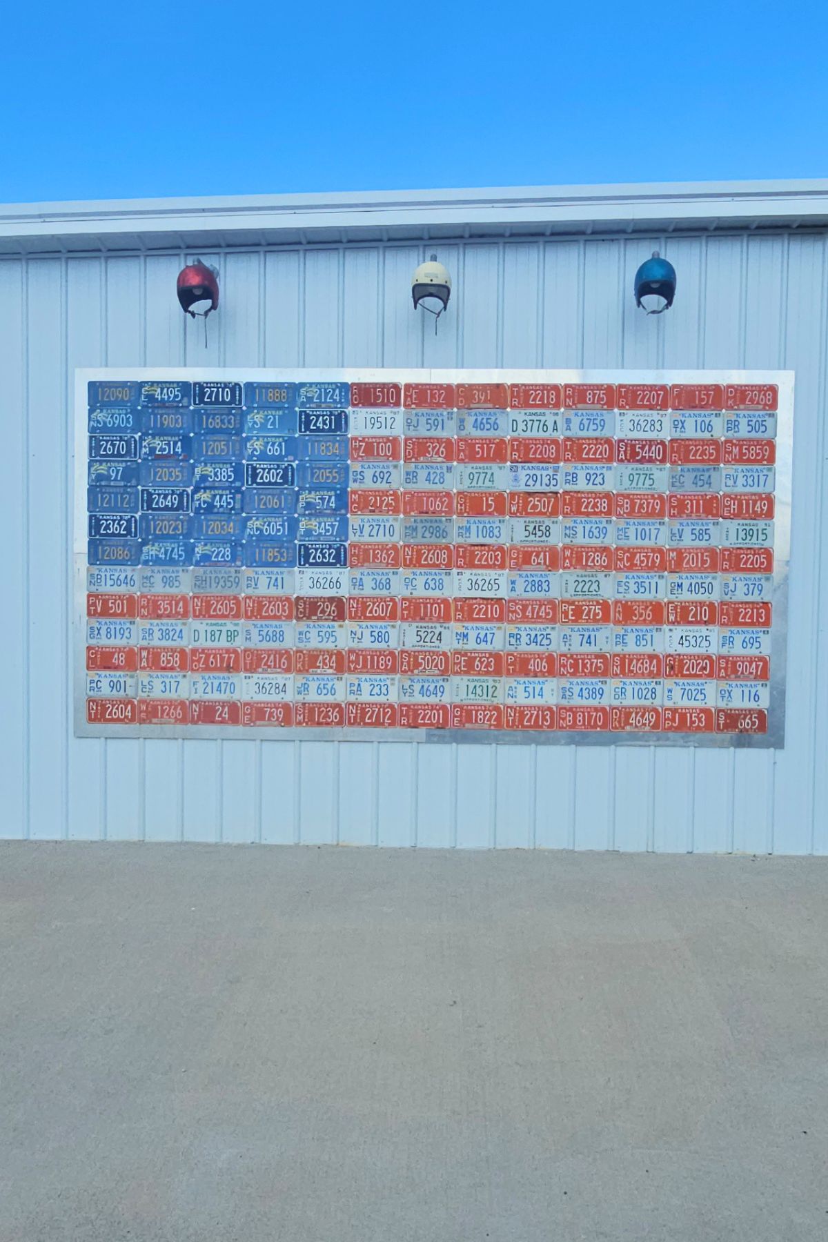 A mural of the American flag made from colored license plates is displayed on the side of a metal building in Southeast Kansas. Three lights are mounted above the mural.