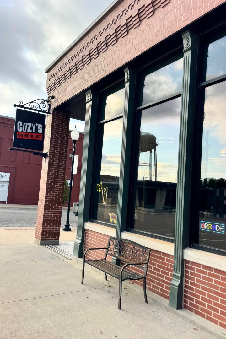 Exterior view of Cozy's Grindhouse in Southeast Kansas with large windows, a bench on the sidewalk, and a water tower reflected in the glass.