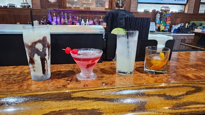 Four assorted cocktails are lined up on a wooden bar counter, capturing the lively spirit of Southeast Kansas, with liquor bottles and a bartender visible in the background.