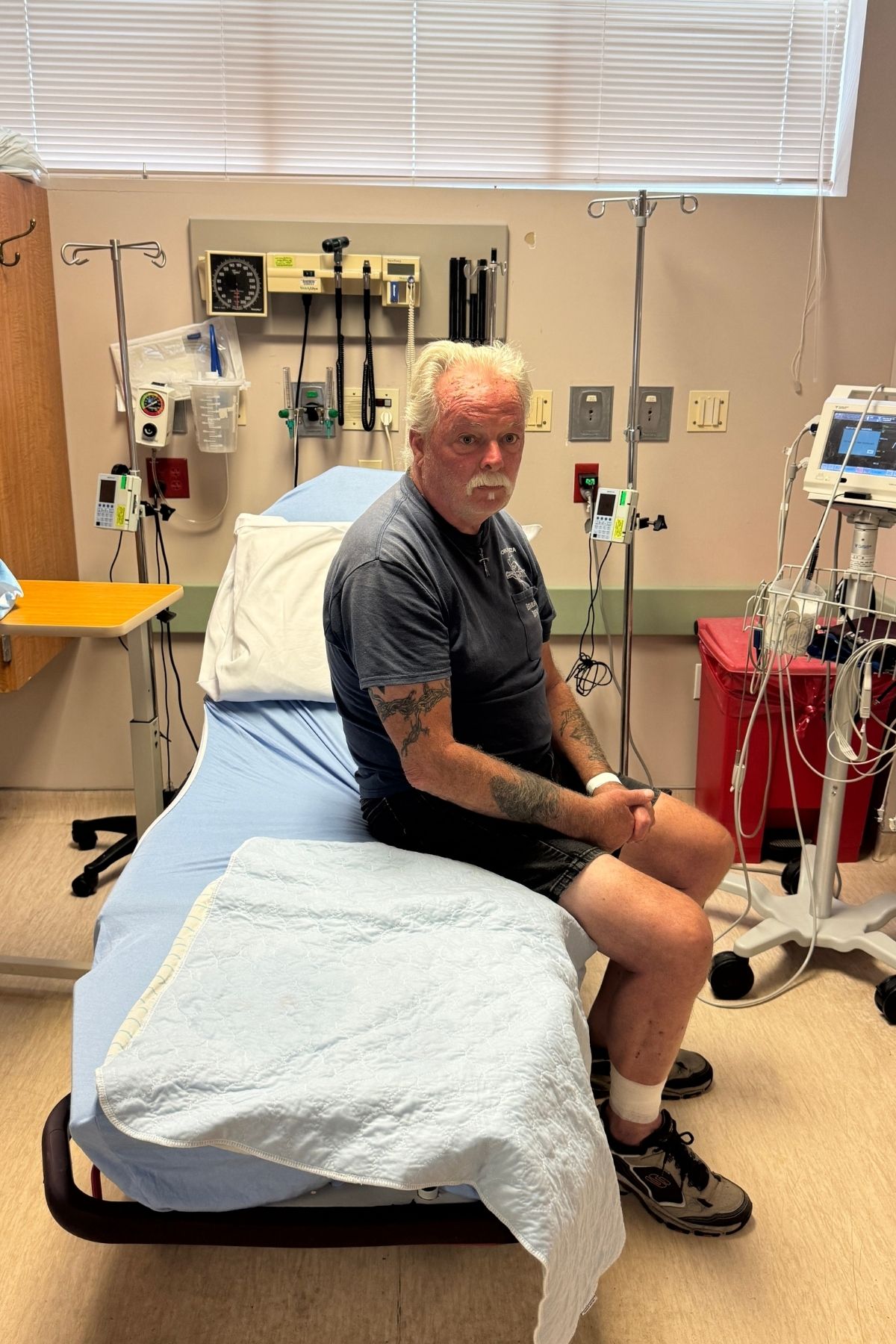 An older man with tattoos sits on a hospital bed in a medical examination room, surrounded by medical equipment and monitors, perhaps pondering life’s big questions—like, “Will You Regret Buying an RV?”.