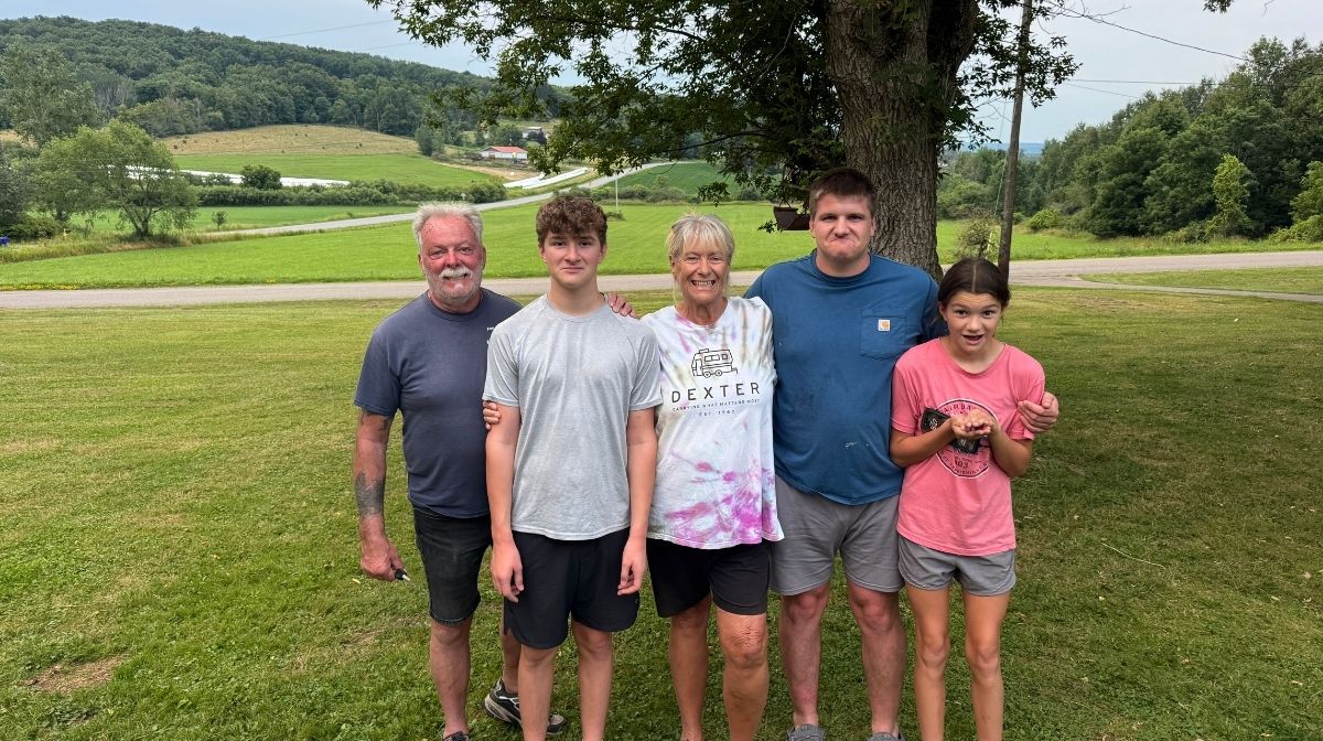 Five people stand together outside on grass with trees, a road, and rolling hills in the background. Two men, one woman, one teenage boy, and one young girl are smiling at the camera—capturing a moment that highlights a key benefit of RV living: the freedom to enjoy beautiful outdoor settings and quality family time wherever you go.