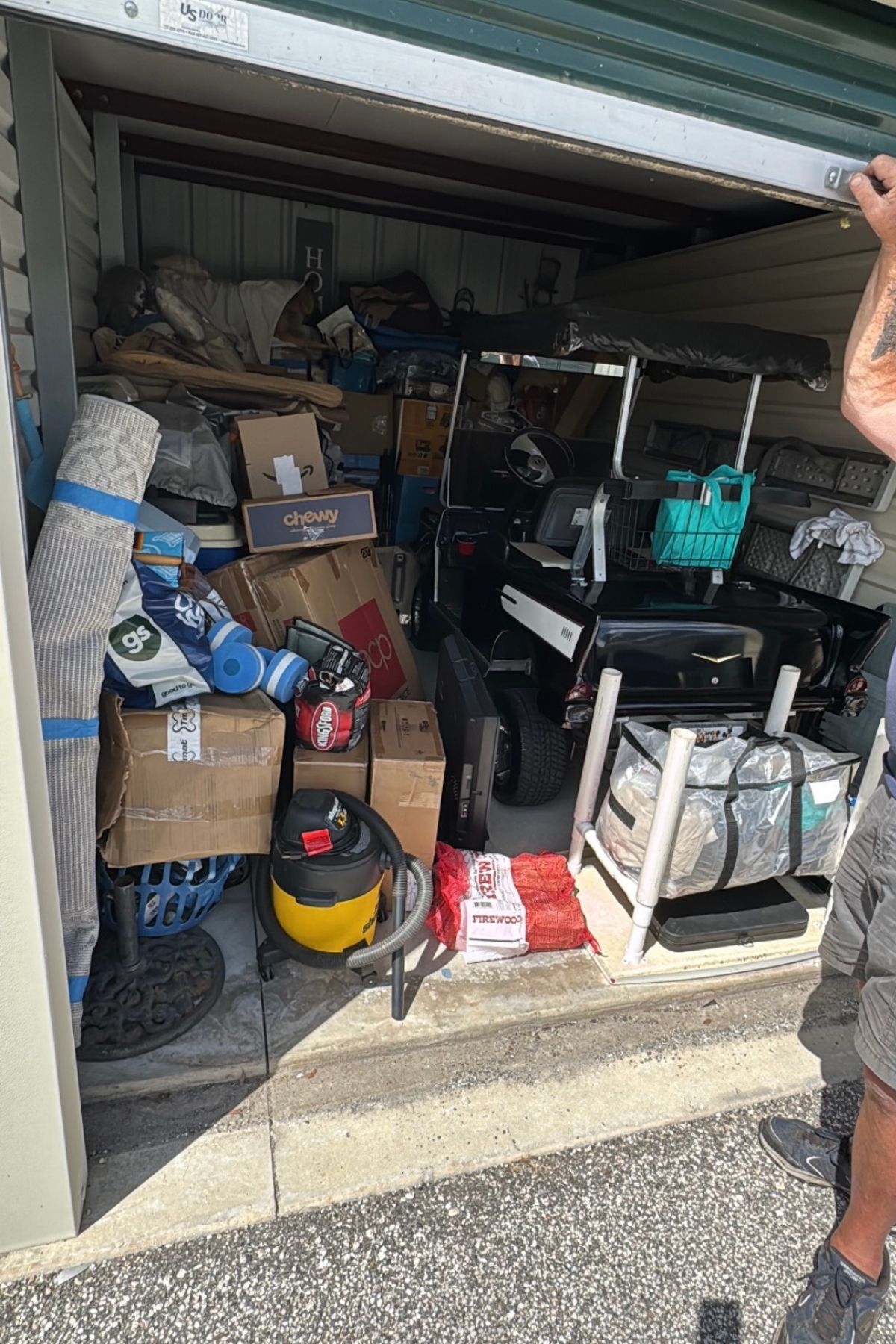 A cluttered storage unit filled with boxes, a golf cart, vacuum, rugs, and various household items hints at the freedom—and occasional challenges—that come with embracing the benefit of RV living. A person is holding the door open on the right side of the image.