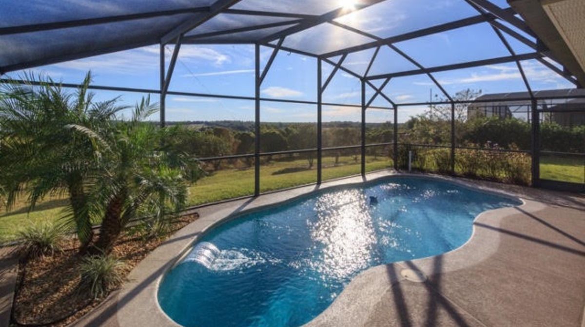 A small backyard pool is enclosed by a screened structure, with a palm tree beside it and a scenic view of a grassy landscape under a blue sky—a relaxing oasis reminiscent of the flexibility and outdoor enjoyment that is a key benefit of RV living.