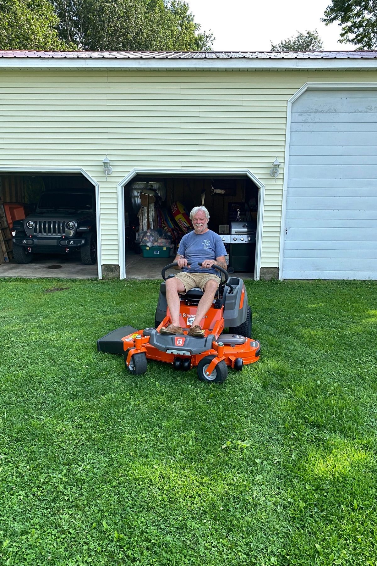 An older man sits on an orange riding lawn mower in front of a garage with two open doors, parked cars, and garden tools visible inside, illustrating the benefit of RV living—having the freedom and space to organize outdoor essentials right at your doorstep.