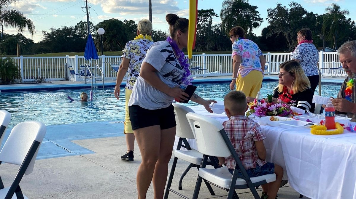 People sit and stand around a table set with food beside an outdoor pool—one child swims, while others talk or serve themselves. The area is decorated for a casual gathering, creating the relaxed atmosphere of America’s new third place.