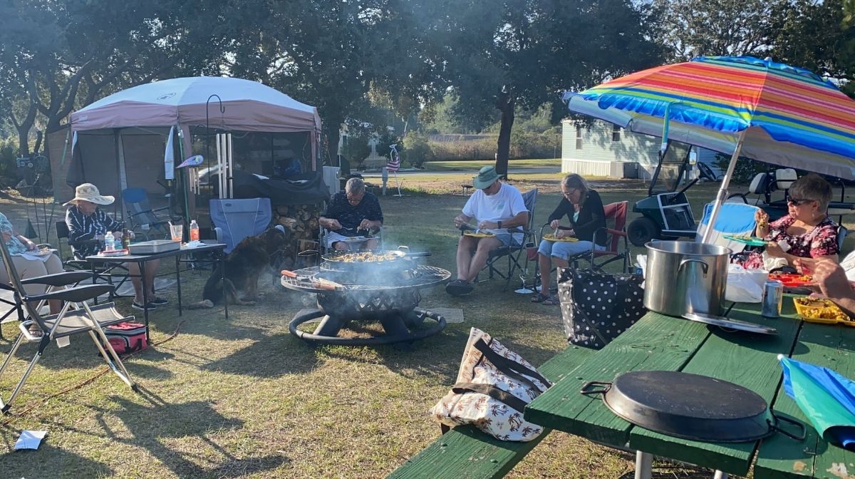 A group of people sit in lawn chairs around a fire pit at a campsite—America’s new third place—with a picnic table, umbrellas, and camping gear visible in the background.
