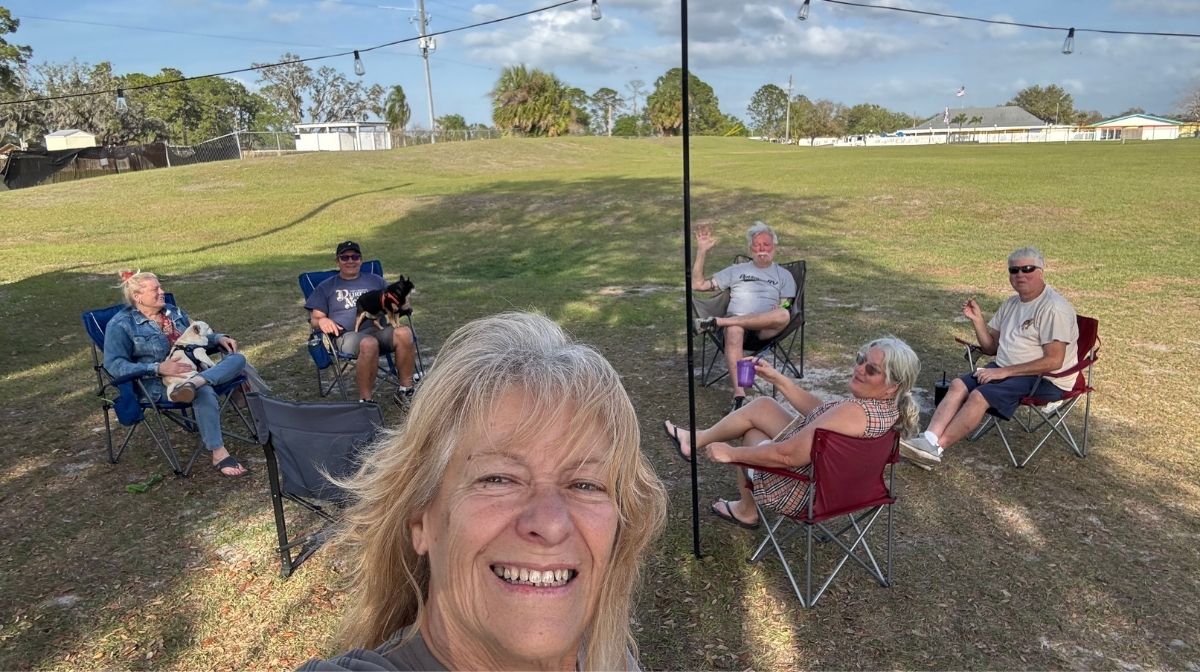 Six adults sit in camping chairs in a grassy field, some waving and smiling at the camera; one holds a small dog. Gathered outdoors on a sunny day, they embody America’s new third place—a welcoming spot for friends, connection, and community beyond home or work.