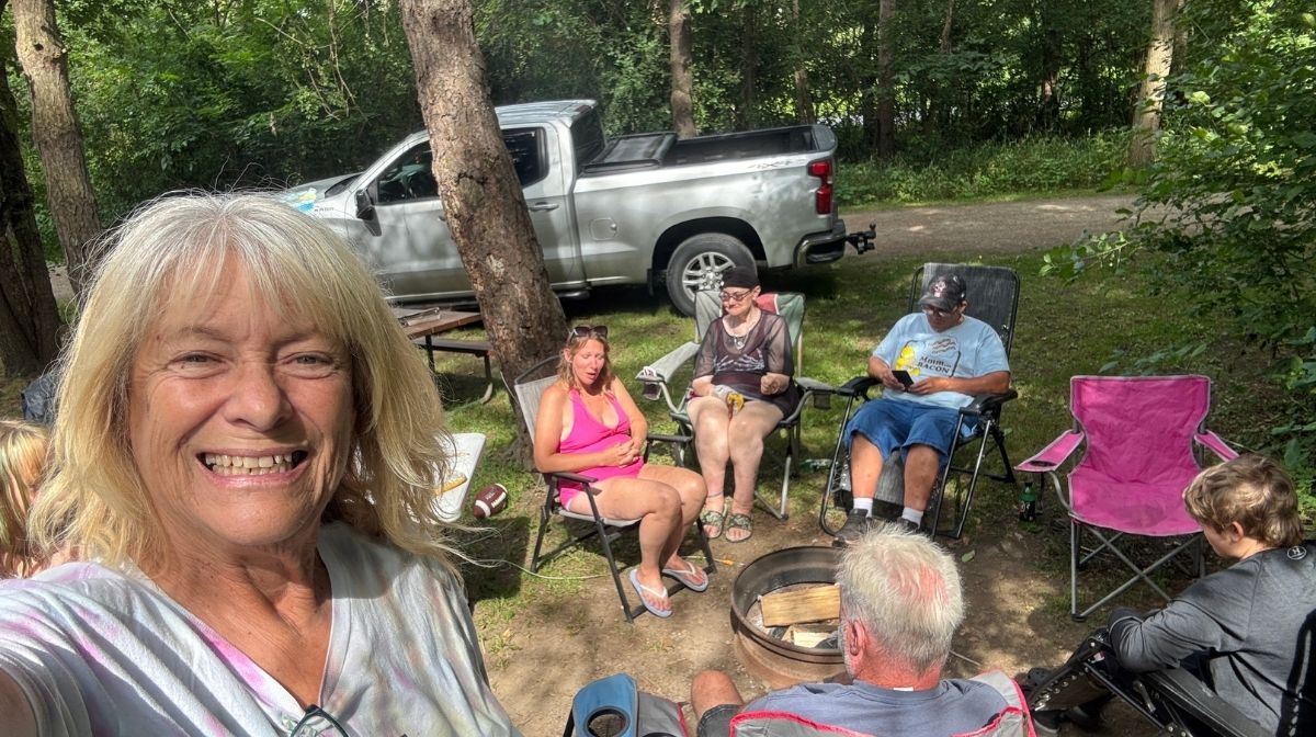A group of people sit around a fire pit outdoors near a parked pickup truck, surrounded by trees—America’s new third place. One woman in the foreground is taking a selfie and smiling at the camera.