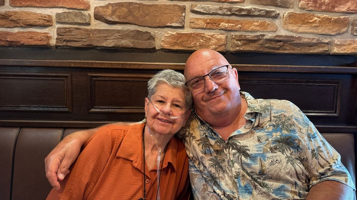 An older woman using oxygen and an older man in a floral shirt sit closely together, smiling, with a brick wall in the background—capturing the warmth and connection found in America’s new third place.