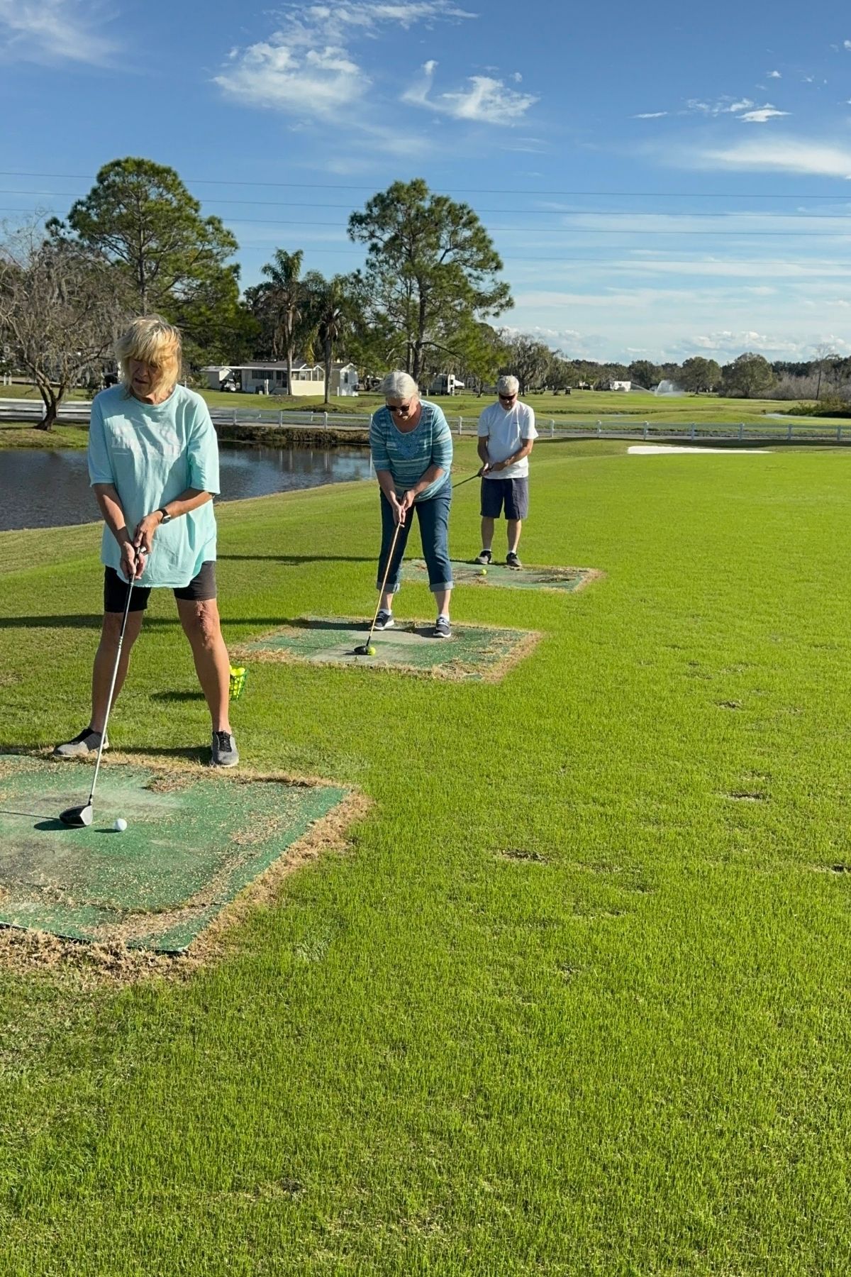 Three people stand in a row on artificial turf mats at a golf driving range, each preparing to hit a golf ball with a club. Trees, water, and blue sky are visible in the background, capturing the laid-back atmosphere of America’s new third place.