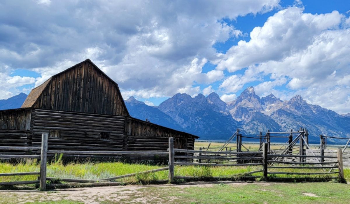 Weathered wooden barn and fence in a grassy field with dramatic mountains and a partly cloudy sky in the background—an iconic scene perfect for your next RV road trip.