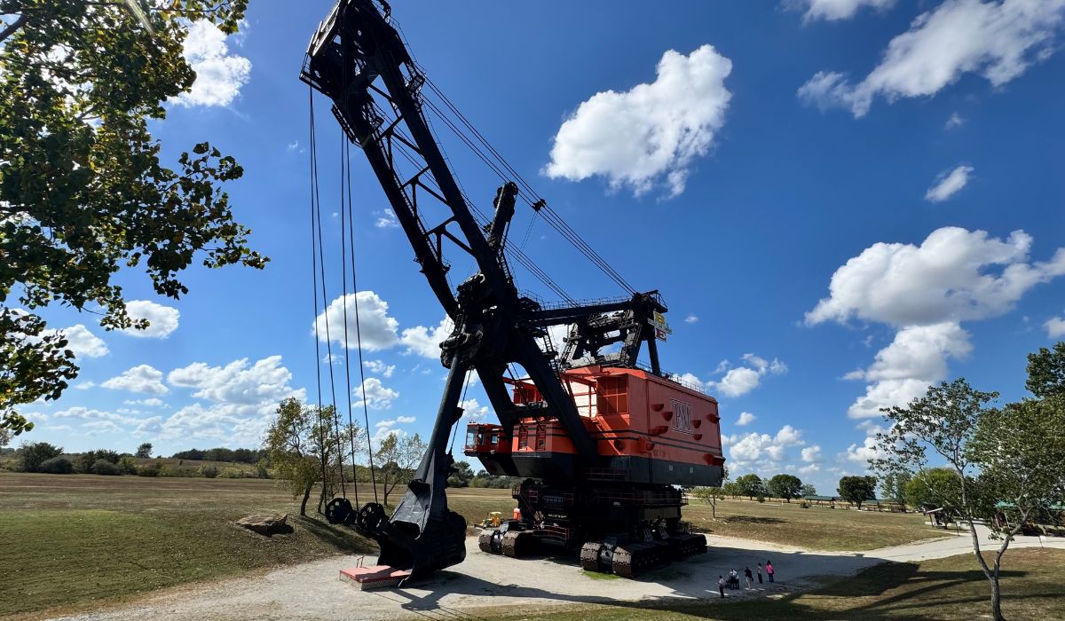 Large black and red mining excavator sits on gravel surrounded by grass and trees under a bright blue sky with scattered clouds; people stand nearby for scale, making it a striking roadside sight often encountered during an adventurous RV road trip.