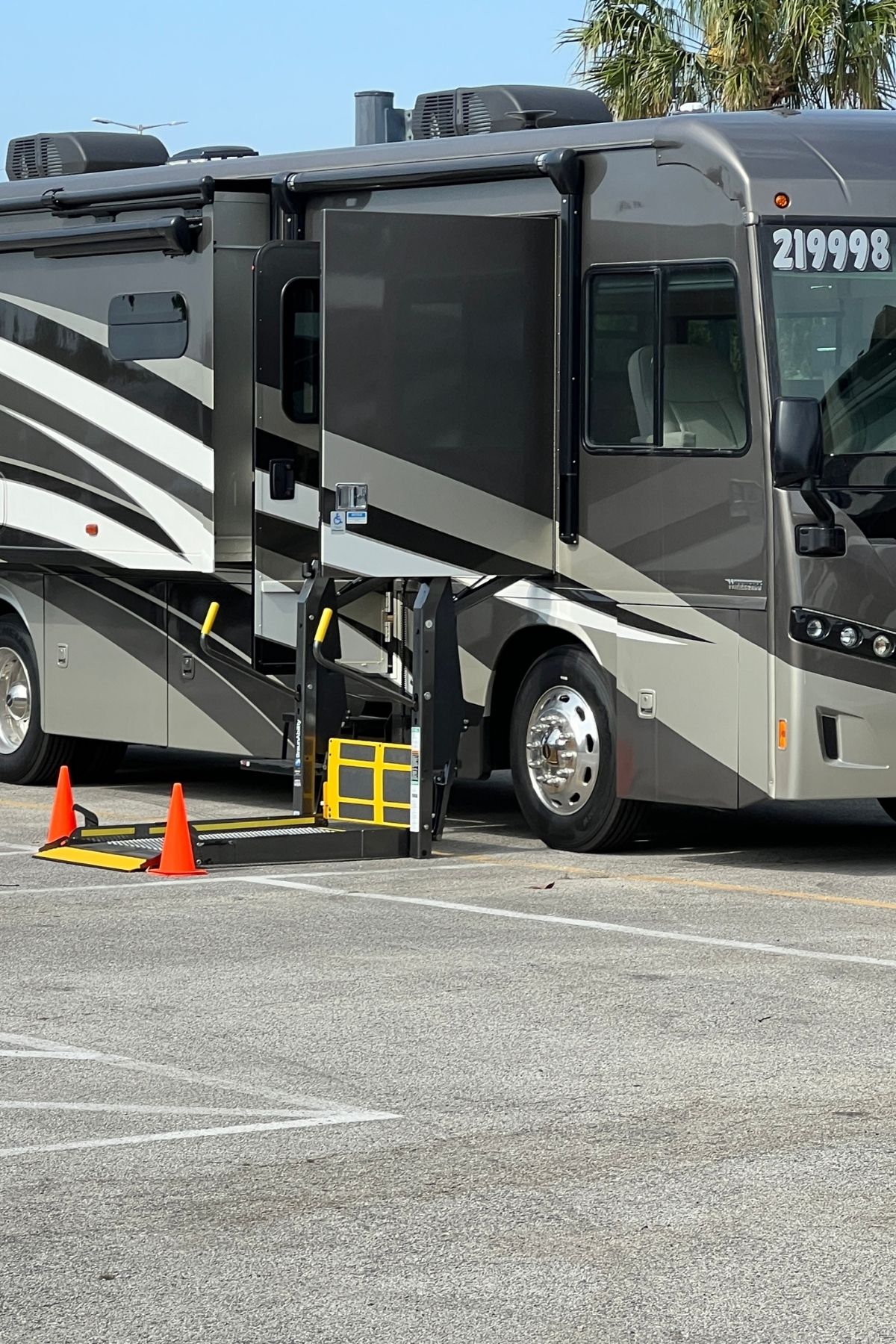 A wheelchair lift is extended from an RV parked in a lot, with orange safety cones placed around the lift—showing just one of the many accessible features you can enjoy when you buy an RV.