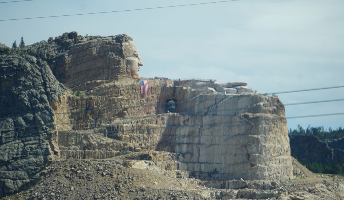 A large rock sculpture resembling a human face and upper body is being carved into a mountainside, with an American flag hanging on the unfinished section—an impressive sight often discovered by travelers during an RV road trip.